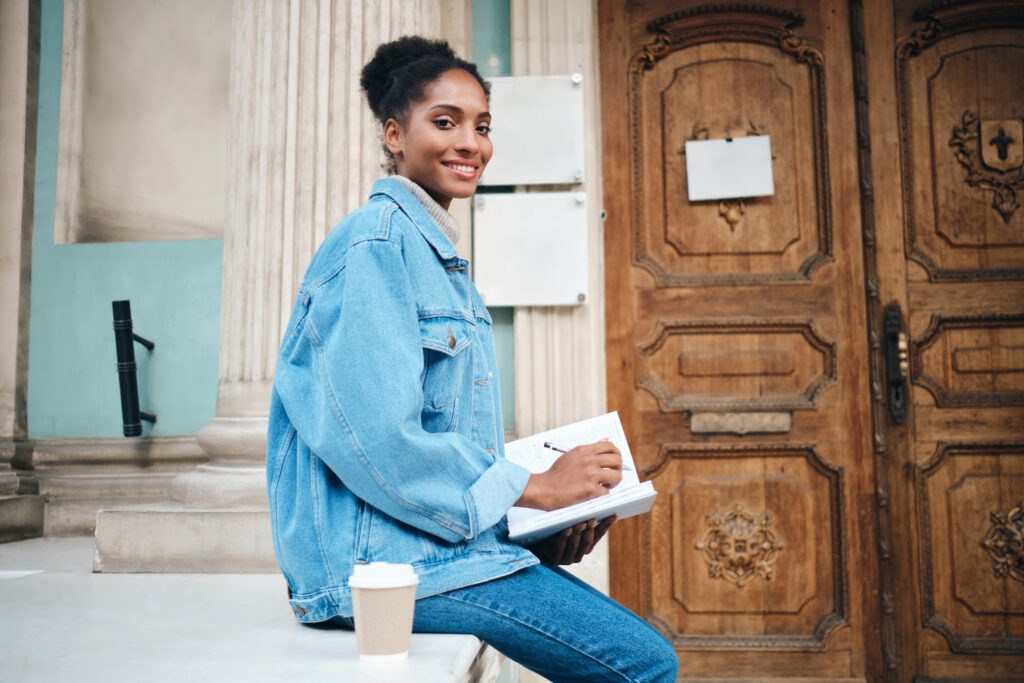 beautiful smiling casual african american student girl in denim jacket with notebook joyfully looking in camera outdoor - Fucape Business School